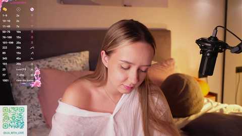 Video of a young, fair-skinned woman with straight, blonde hair, wearing a white off-shoulder top, sitting on a bed with floral bedding, using a microphone, in a cozy bedroom.
