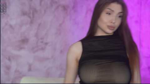 Video of a young, fair-skinned Asian woman with long, straight black hair, wearing a black, sheer top revealing her ample breasts. She stands against a textured, marbled purple wall.