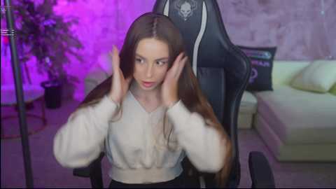 Video of a young woman with long brown hair, wearing a white blouse, adjusting her hair in a gaming chair with a skull logo. Background includes a purple-lit room with a white sofa, potted plant, and brick wall.