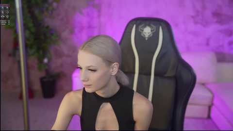 Video of a slender, light-skinned woman with blonde hair in a high ponytail, wearing a black sleeveless top with a keyhole neckline, seated in a black gaming chair with purple ambient lighting in the background.
