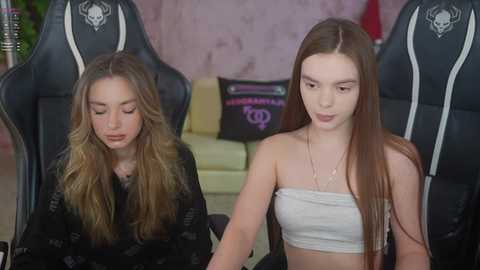 Video of two young women in a gaming room, one with long brown hair, the other with long blonde hair, sitting in black gaming chairs.