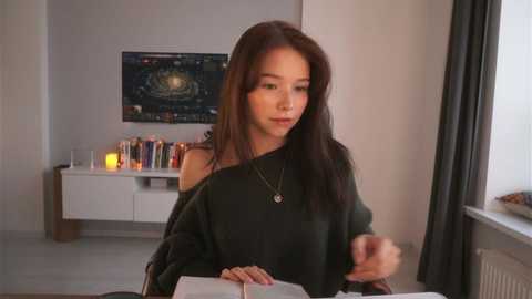 Video of a young woman with long brown hair, wearing a dark sweater, sitting at a table in a modern, minimalist living room with a large window, a white shelf, and a colorful painting on the wall.