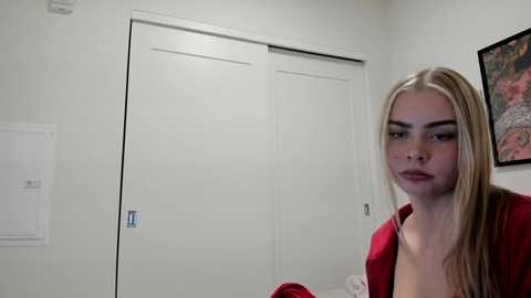 Video of a young woman with long blonde hair, fair skin, and red lipstick, wearing a red blouse, standing in a minimalist bedroom with white walls, a closed white door, and a colorful painting on the wall.