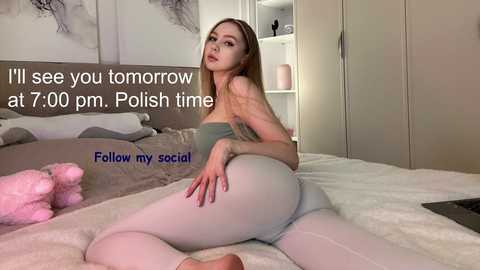 Media: A video of a young woman with fair skin and long blonde hair, wearing a strapless top and tight white leggings, sitting on a bed in a bedroom. Text overlay reads, \"I'll see you tomorrow at 7:00 pm, Polish time.\