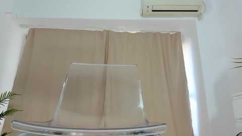 Video of a clear acrylic chair with a minimalist design, positioned in front of beige curtains, against a white wall with an air conditioning unit mounted above.
