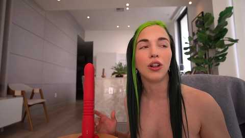 Video of a fair-skinned, topless woman with green streaked hair, holding a red dildo, in a modern, minimalist living room with plants and wooden furniture.