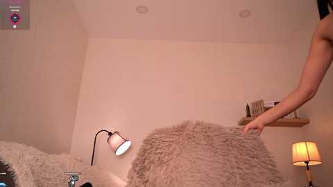 Video of a person in a cozy bedroom, adjusting a fluffy beige throw on a bed. The room has a minimalistic, warm ambiance with a wooden lampshade, a small shelf, and recessed ceiling lights.