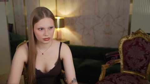 A video of a young woman with long, straight blonde hair, fair skin, and wearing a black lace camisole, sitting in a dimly lit, ornate room with a green sofa and a gold-patterned chair.