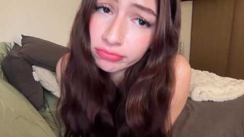 Video of a young woman with long, wavy brown hair and fair skin, wearing a nose ring, posing on a bed with green and brown sheets, giving a slight pout.
