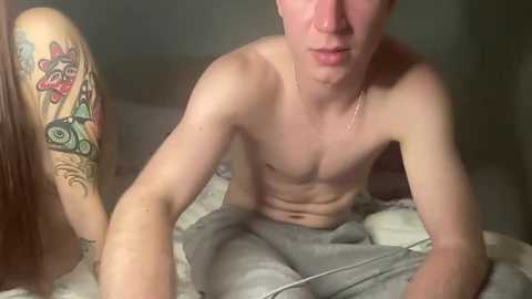 Video of a shirtless, slender white man with short hair, seated on a bed. He has a tattoo of a stylized face on his upper arm. The background shows a blurred, dimly lit room with a bed covered in white sheets.