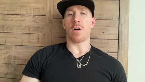 Video of a Caucasian man with a beard, wearing a black baseball cap, black t-shirt, and multiple dog tags, standing against a wooden paneled wall.