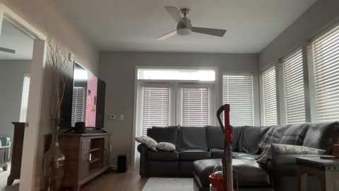 Video of a modern, minimalist living room with a dark grey sectional sofa, flat-screen TV on a wooden stand, and large windows with white blinds. Ceiling fan above.