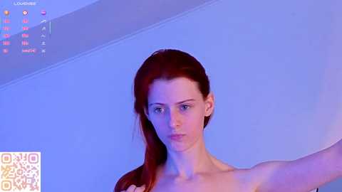 A video of a fair-skinned woman with red hair, wearing a white top, standing against a blue background. QR code at the bottom left corner.