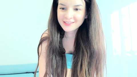 Video of a young Caucasian woman with long, straight brown hair, wearing a blue strapless top, smiling. The background features a light blue wall and a window with vertical blinds, suggesting an indoor setting.