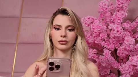 Media: Video of a fair-skinned, blonde woman with long hair, wearing pink eyeshadow and pink lipstick, holding a phone, against a pink background with cherry blossom trees.