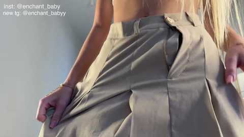 Video of a blonde woman's back, wearing a beige pleated skirt, lifting it to reveal her underwear.