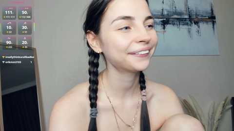 Video of a young woman with long, braided black hair, wearing a black tank top, smiling softly, with a modern, abstract painting in the background.