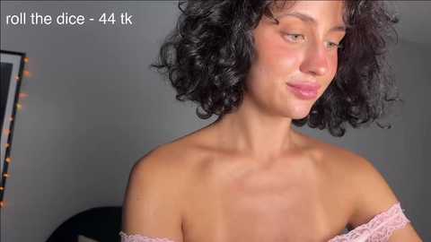 Video of a young woman with curly black hair, fair skin, and light makeup, wearing a pink lace off-shoulder top. She's smiling subtly. Text overlay reads \"roll the dice - 44k.\