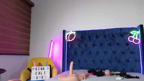 Video of a bedroom with a tufted blue velvet headboard, neon lights, a pink dildo on a yellow chair, and a sign reading \"Gina Cali XXX.\