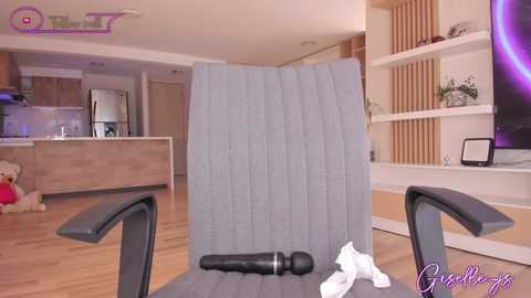 Video of a modern, minimalist living room featuring a gray chair with a black microphone and a white tissue, wooden floors, white shelves, and a flat-screen TV mounted on the wall.
