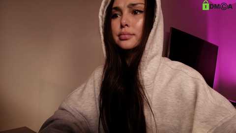 Video of a woman with long dark hair, wearing a gray hoodie, sitting indoors with a beige wall and purple lighting in the background.