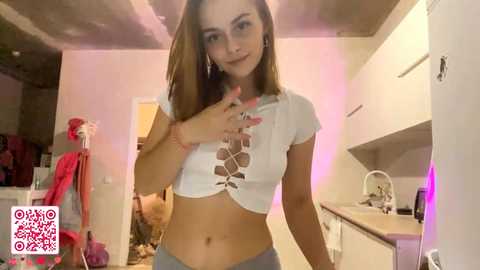 Video of a young Caucasian woman with light skin, straight brown hair, and a slender physique, wearing a white crop top with lace detailing and gray pants, posing in a modern kitchen with white cabinets and pink accents.