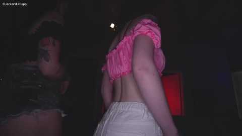 Video of a woman with light skin, wearing a pink ruffled crop top and beige pants, captured from behind, with a dimly lit, shadowy background.