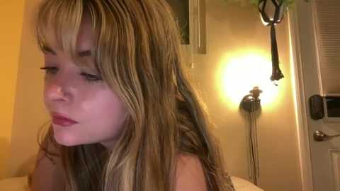 Video of a young woman with long, wavy blonde hair, fair skin, and a contemplative expression, in a dimly-lit bathroom with a yellow wall and a towel rack.