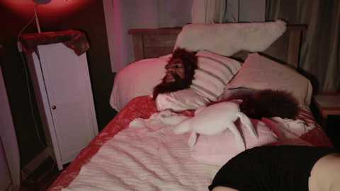 Video of a man with a bushy beard and wild hair, lying on a bed in a dimly lit room with red lighting. He is covered in a pink blanket, with a white dog resting next to him.