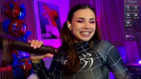 Video of a smiling, light-skinned woman with long brown hair, wearing a black Spider-Man suit, holding a large, dark dildo, in a dimly-lit room with purple lighting, framed by decorative objects and a TV screen displaying streaming content.