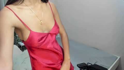 Video of a light-skinned woman in a pink satin camisole, revealing her small breasts and nipples, with a heart pendant necklace, in a minimalist bedroom with a bed and a phone.