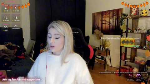 Video of a young blonde woman in a white robe, eyes closed, in a cozy, cluttered bedroom with gaming and personal items; streaming overlay visible.