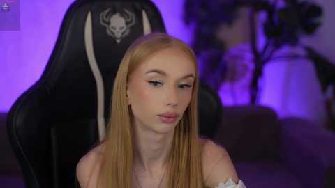 Video of a young, fair-skinned, blonde woman with long, straight hair, wearing a white off-shoulder top, sitting in a black gaming chair with a skull emblem, in a dimly lit room with purple ambient lighting.