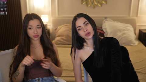 Video of two young women, one with long brown hair, wearing a black top, and the other with long black hair, wearing a black velvet top, sitting on a white couch in a modern, dimly lit bedroom.
