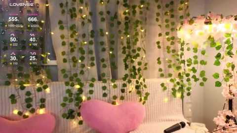 Video of a cozy bedroom with a white radiator, green ivy garlands, pink heart-shaped pillows, and warm fairy lights.
