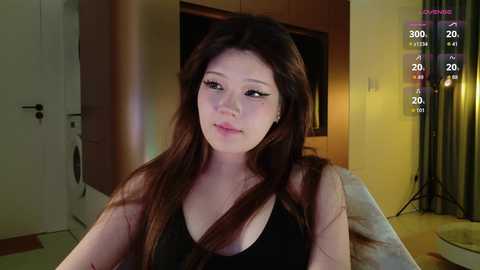 Video of an Asian woman with long, dark hair in a black tank top, indoors with dim lighting, smiling slightly.