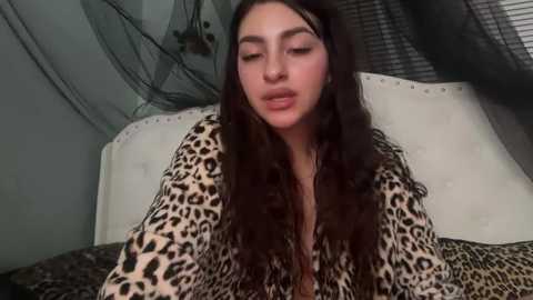 Video of a young woman with long, wavy dark hair, wearing a leopard-print robe, sitting on a bed with white headboard and dark curtains.