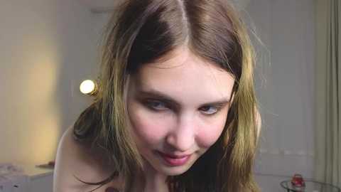 Video of a young, fair-skinned woman with long, wavy brown hair, slightly parted lips, and a soft, slightly mischievous expression. Background shows a softly lit room with white walls and a blurry table lamp.