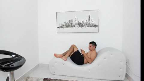 Video of a shirtless, muscular man with short hair, wearing black shorts, lounging on a white chaise in a minimalist room with a black chair and a cityscape artwork on the wall.