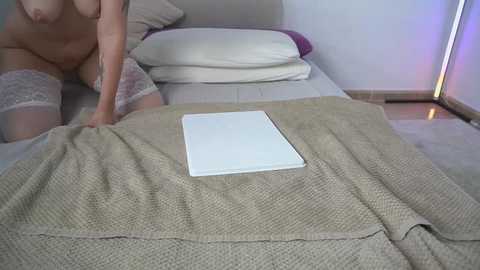 A video of a nude woman with fair skin, wearing only thigh-high lace stockings, pulling a beige blanket off a bed with white sheets.