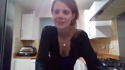 A video of a smiling Caucasian woman with light skin and shoulder-length brown hair, wearing a black top, in a modern kitchen with white cabinets and stainless steel appliances.