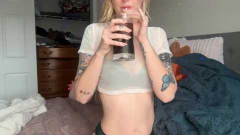 Video of a blonde, slim, tattooed woman in a sheer, white crop top, drinking from a glass in a messy bedroom with a bed, dresser, and door.