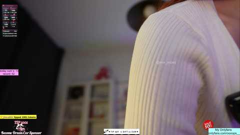 A video of a woman in a white ribbed sweater, captured from a video game stream, with a blurred background featuring a bookshelf and a dark wardrobe.