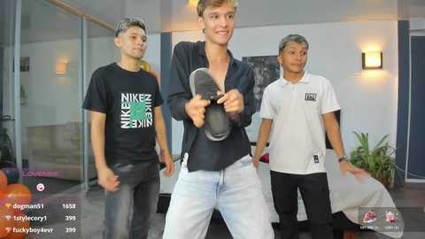 Video of a young man in a black shirt, holding a microphone, surrounded by two men in casual clothes in a modern, indoor setting.