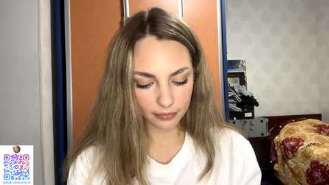 A video of a young woman with long, straight, light brown hair, wearing white clothing, with closed eyes and serene expression, in a room with orange and blue cabinets, a floral bedspread, and a QR code in the lower left corner.