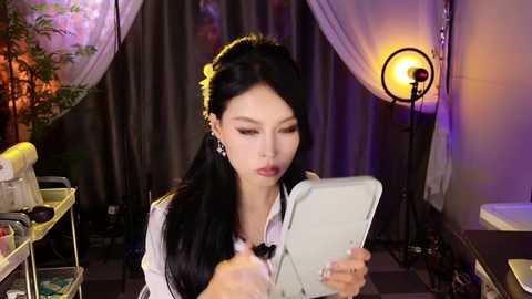 Video of a woman with long black hair, fair skin, and makeup, using a makeup brush in a dimly lit, well-lit vanity room with purple lighting, framed mirrors, and a large plant.