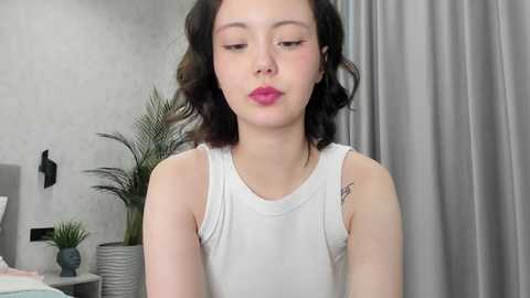 A video of a young Asian woman with fair skin, wavy black hair, and pink lips, wearing a white sleeveless top, sitting in a modern, light-colored room with a potted plant and gray curtains.
