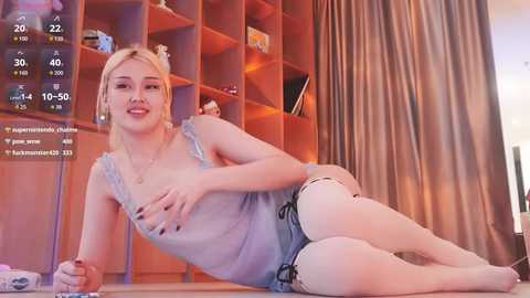 A video of a blonde woman with fair skin, wearing a sheer blue top and denim shorts, lying on a wooden floor, surrounded by a modern bookshelf and curtains.