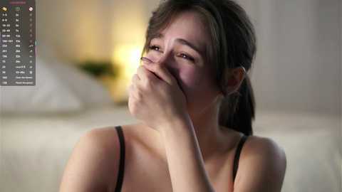 A video of a young Asian woman with long brown hair, wearing a black spaghetti-strap top, covering her mouth with her hand, crying, blurred background, soft lighting.
