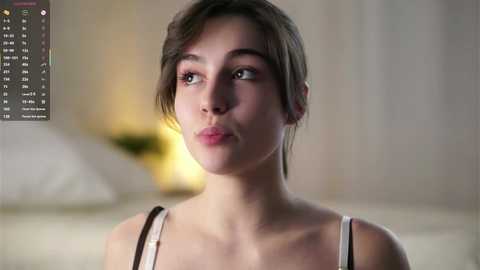 A video of a young woman with light skin, brown hair, and light makeup, wearing a white tank top, pensive expression, blurred background.